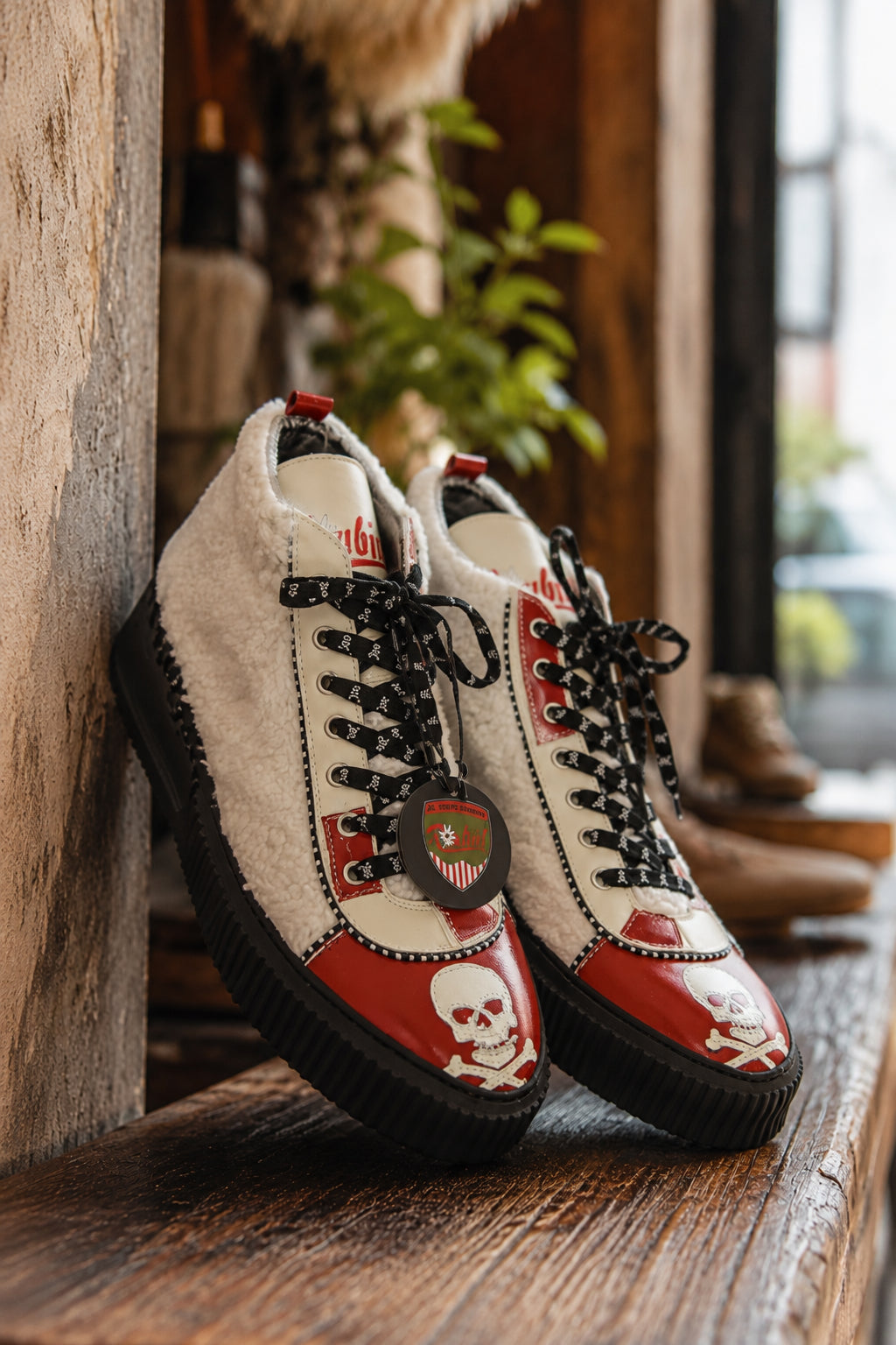 Rot-weiße Raubirl Lederschuhe mit Fellbesatz, Totenkopf-Motiv und schwarzen Schnürsenkeln im rustikalen Werkstatt-Ambiente